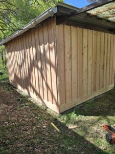 Bardage vertical en pin clair sur un abri extérieur. Base renforcée et gouttière installée. Ombres de feuillage sur le mur.