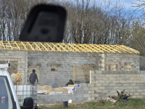 Chantier de construction avec murs en blocs et charpente bois jaune fraîchement posée. Deux ouvriers travaillent près du sable et d'une bétonnière. Marque "Alumar" sur le mur.