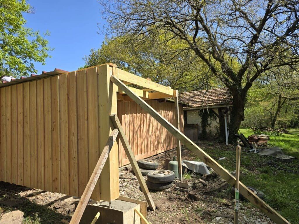 Chantier extérieur. Poteau d'angle en bois fixé sur plot béton, étais de soutien, et bardage vertical d'un nouvel abri sous un ciel bleu.