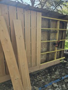 Rénovation d'une cabane: pose de bardage bois vertical sur une ossature jaune. Planches en attente. Un cloueur et un niveau sont visibles.