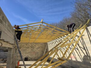 Deux charpentiers fixent une charpente préfabriquée en bois (fermettes) sur des murs de blocs, utilisant des outils, sous un ciel bleu.