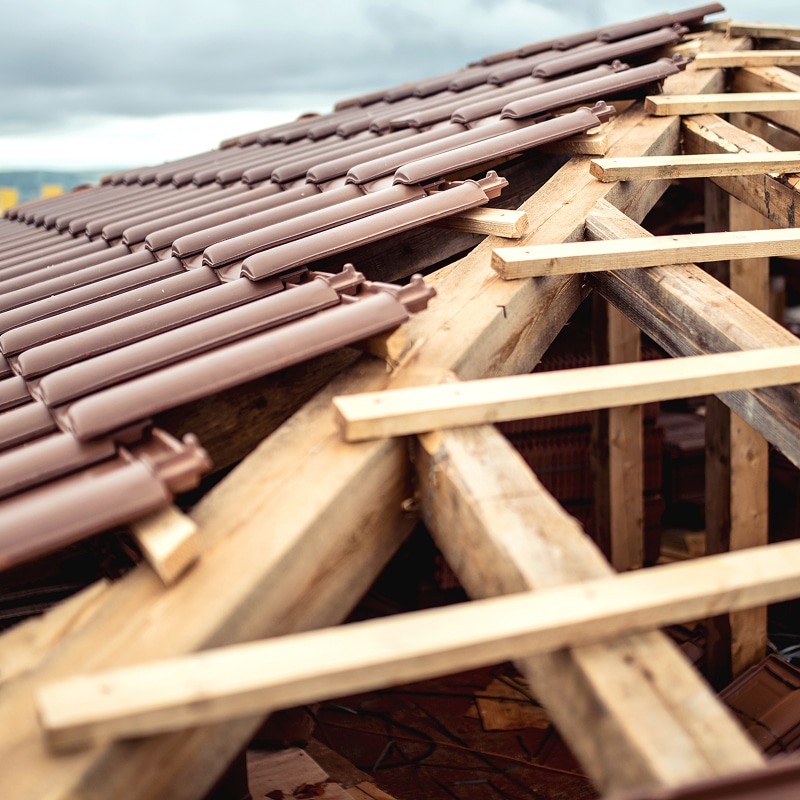 Toiture en construction avec des tuiles marron en cours de pose sur une charpente en bois robuste sous un ciel nuageux.