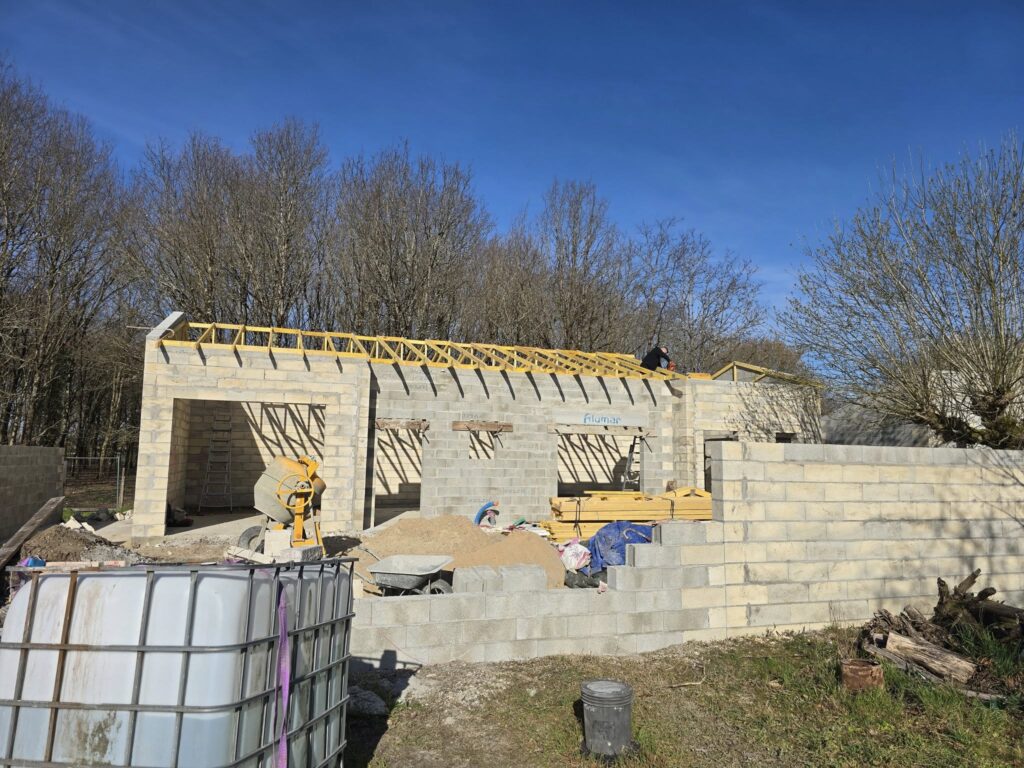 Construction d'une maison en parpaings sous ciel bleu. Charpente bois en place, ouvrier sur le toit. Bétonnière, citernes et sable au sol. Alumar.