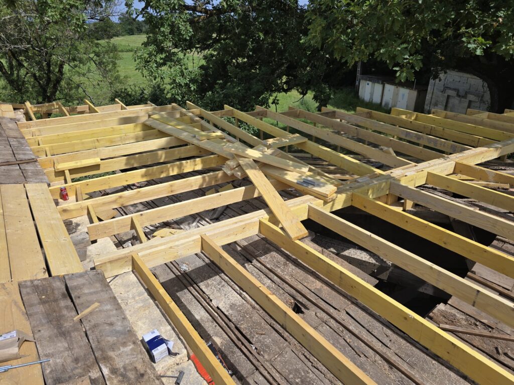 Structure de charpente en bois clair en construction sur un ancien plancher, avec vue sur un champ verdoyant et des arbres.