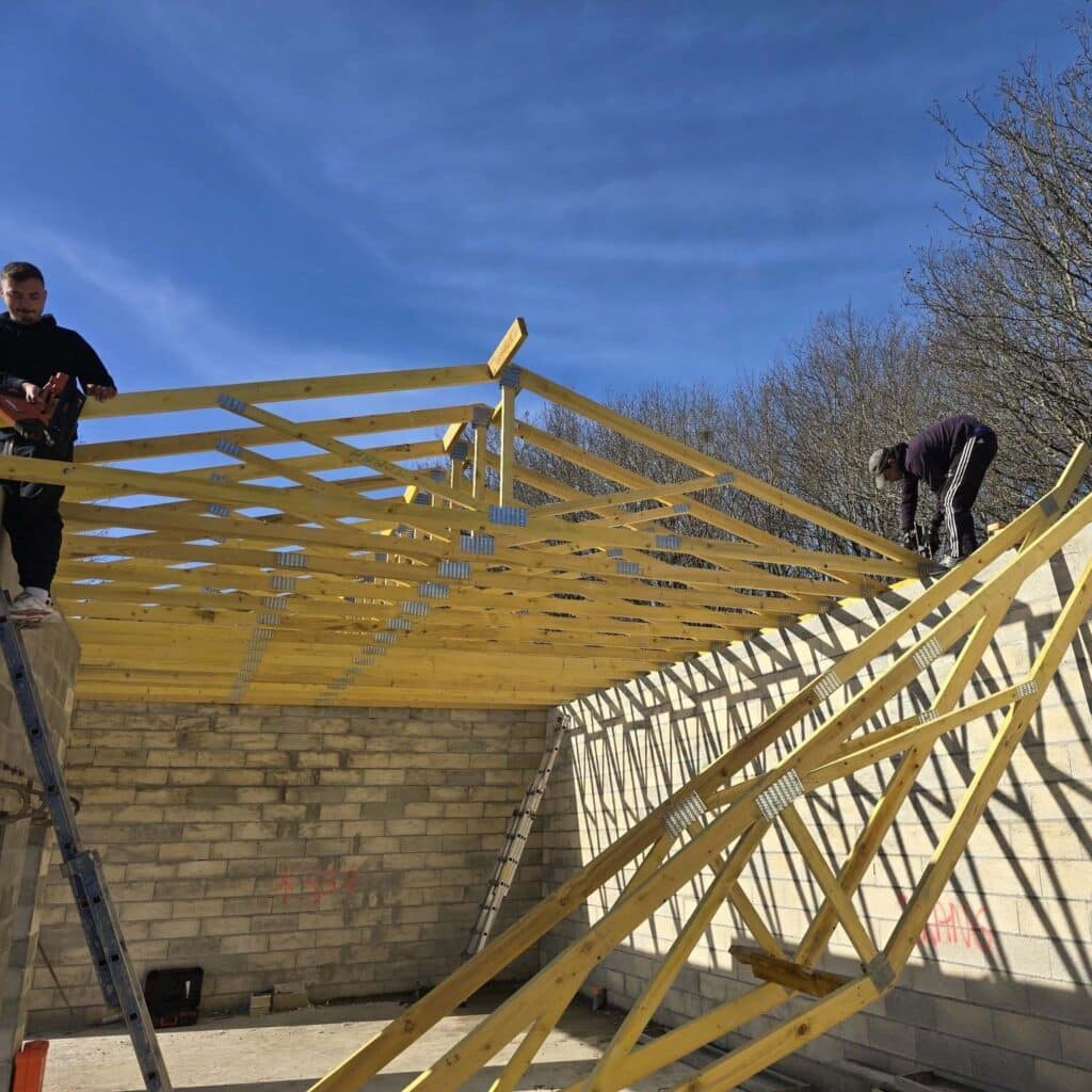 Deux ouvriers construisent une charpente de toit en bois sur un bâtiment en maçonnerie, sous un ciel bleu. Le texte 'X3-1' est visible sur le mur.