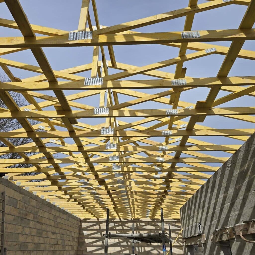 Charpente de toit en bois neuve avec solives et fermes, assemblées par des connecteurs métalliques, sous un ciel bleu dégagé.