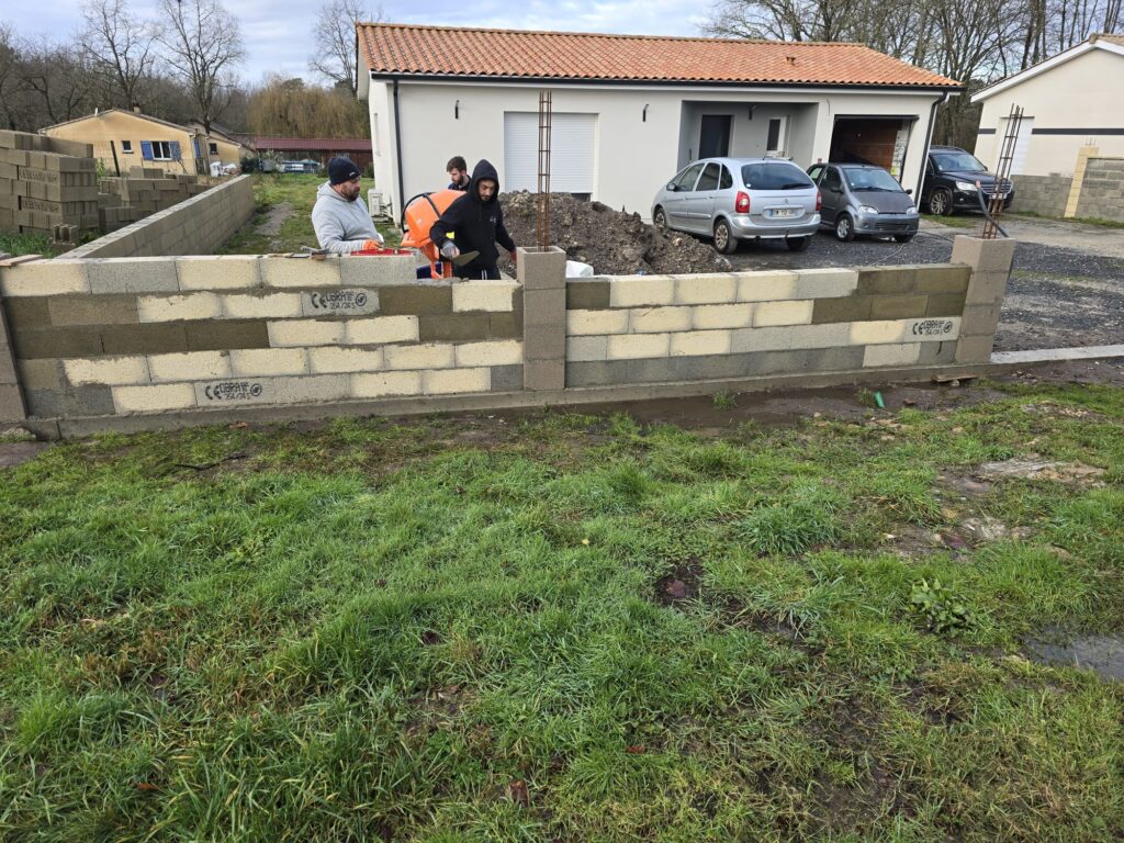Trois maçons posent des parpaings OBRA® CE 20/20 M avec une bétonnière, érigeant un mur devant une maison moderne et des voitures.