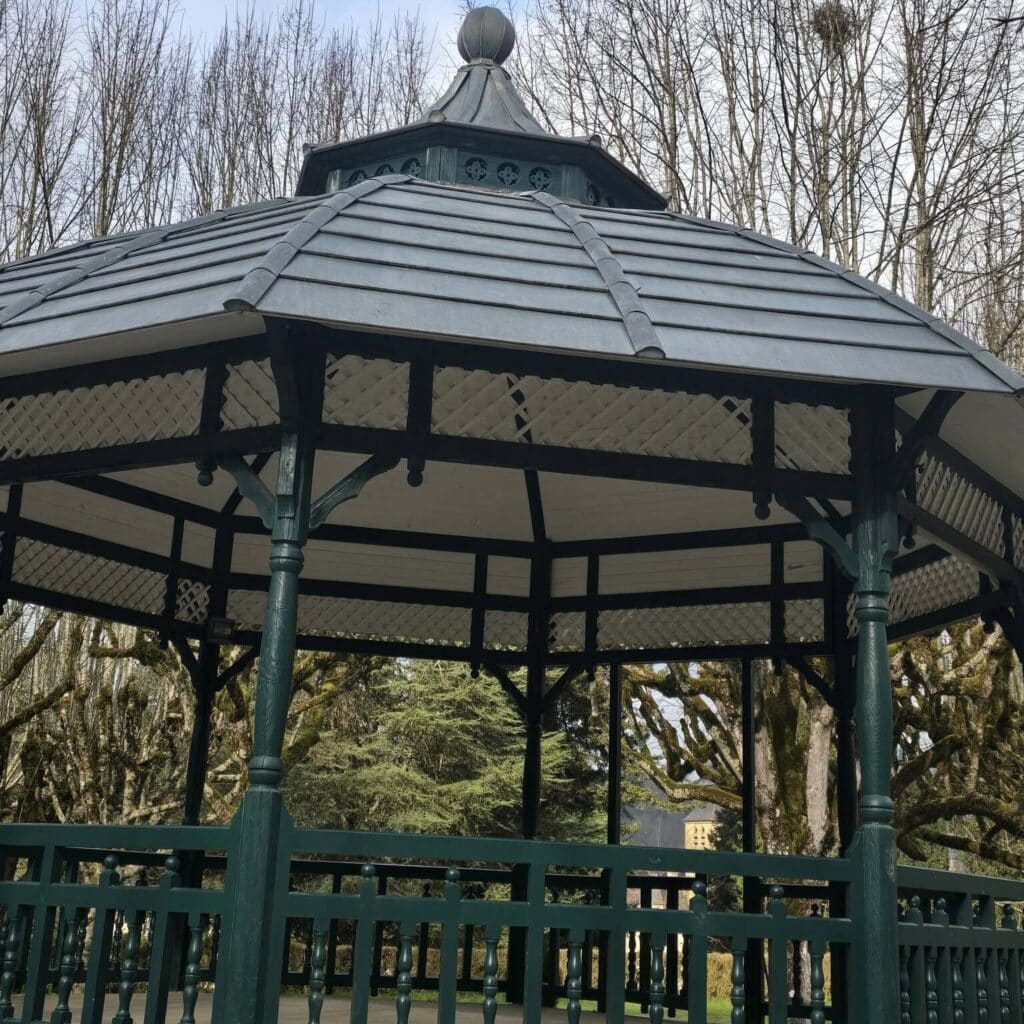 Gazebo vert foncé avec toit gris, lattis décoratif. Entouré d'arbres nus, certains moussus, et un bâtiment au loin.