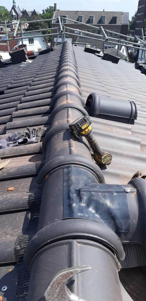 Rénovation de toiture en tuiles sombres avec conduit de ventilation noir et échafaudage en arrière-plan. Lampe de travail posée sur le conduit.