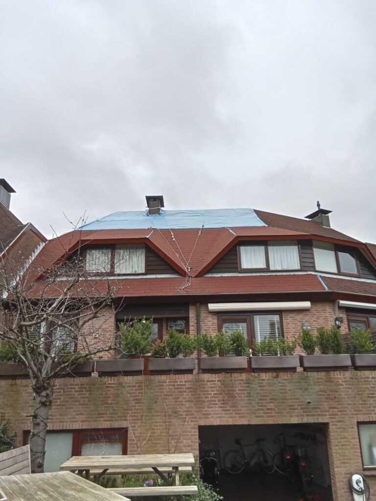 Grande maison au toit de tuiles rouges, une bâche bleue couvre une section pour réparation. Ciel nuageux, arbres au premier plan.
