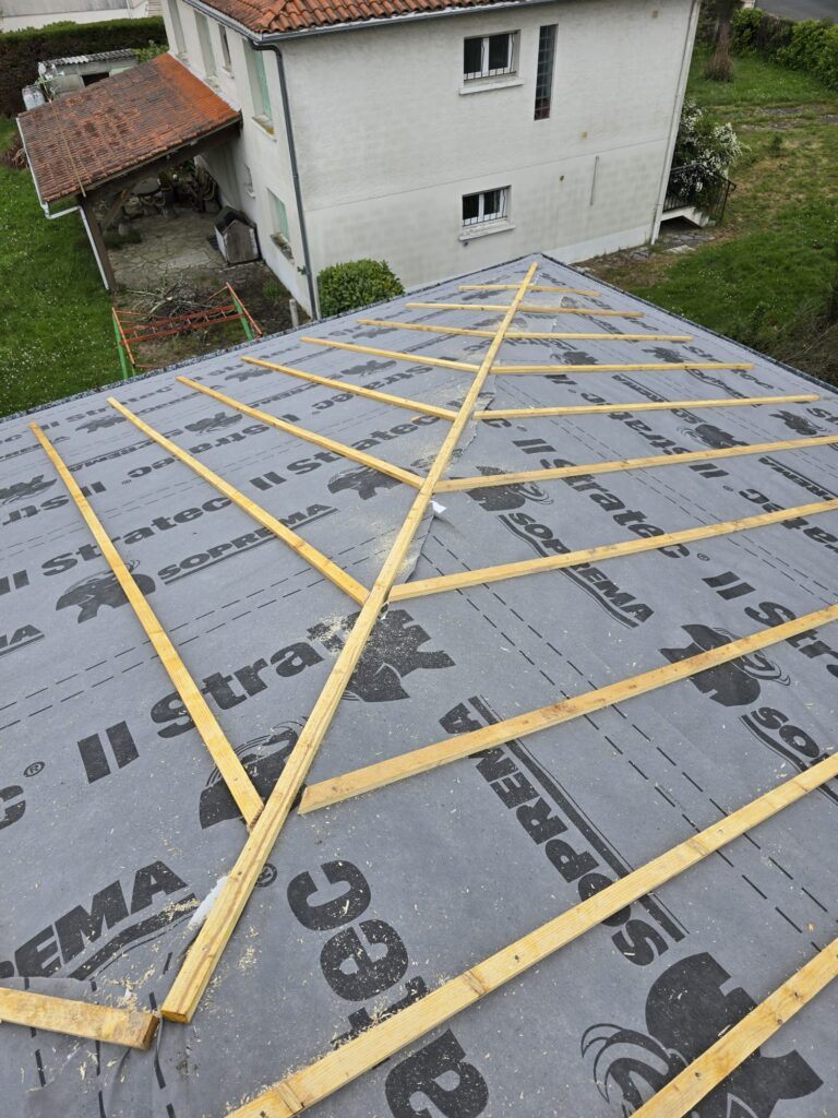 Toiture en construction avec membrane d'étanchéité SOPREMA Stratec grise, recouverte de lattes en bois. Sciure fraîche. Maison en arrière-plan.