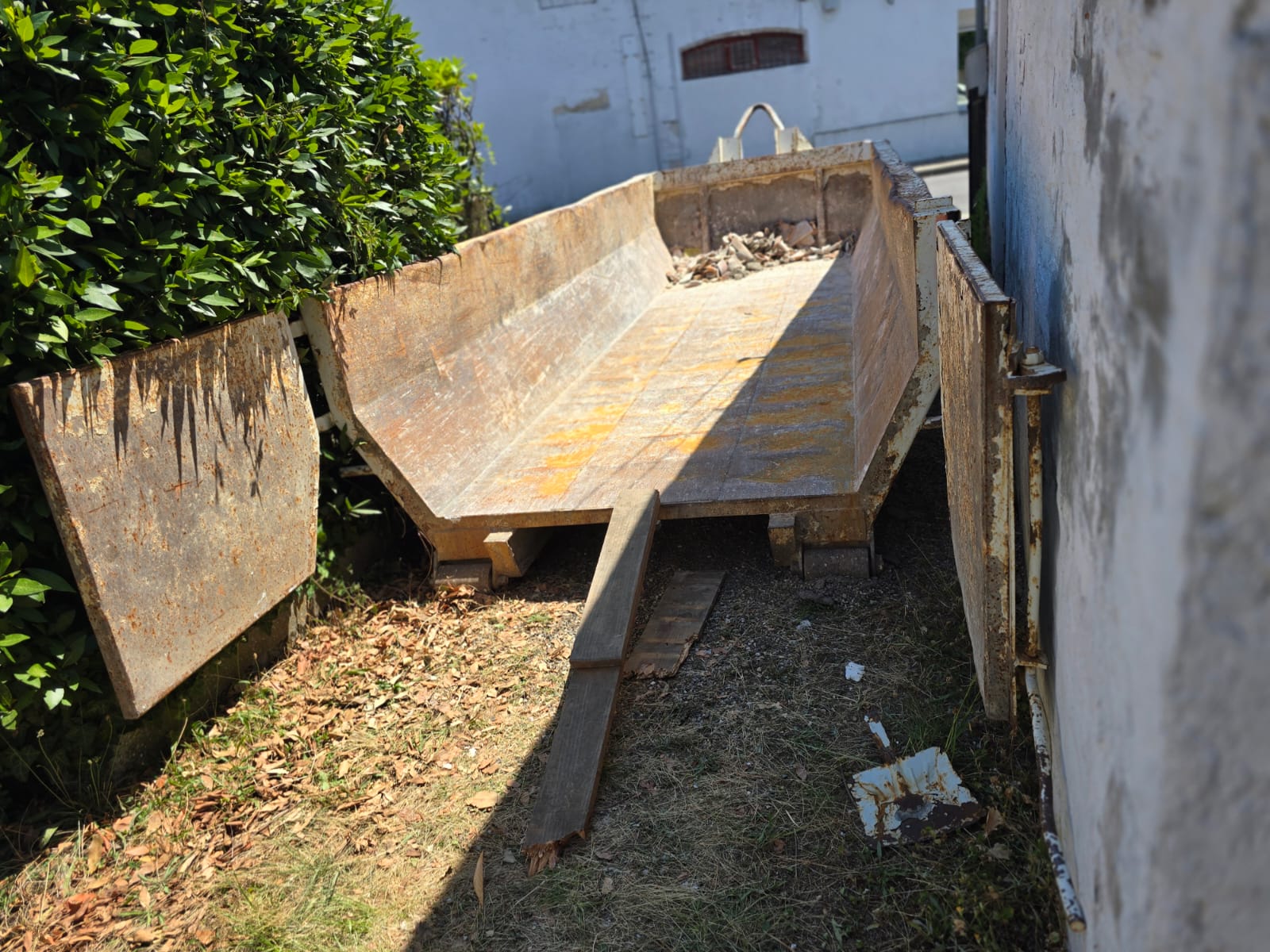 Benne à gravats en métal rouillé ouverte sur un terrain extérieur entre une haie et un mur de bâtiment.