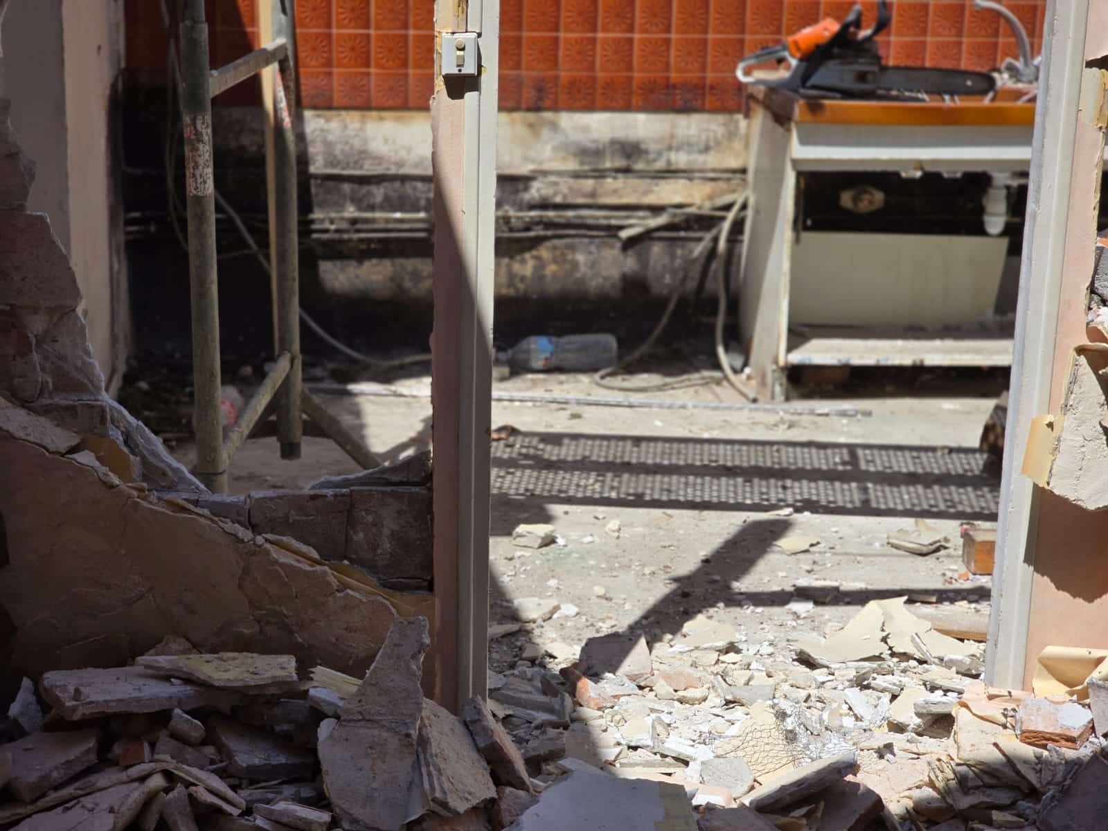 Chantier de démolition avec gravats au sol, échafaudage et une tronçonneuse posée sur un établi devant des carreaux orange.