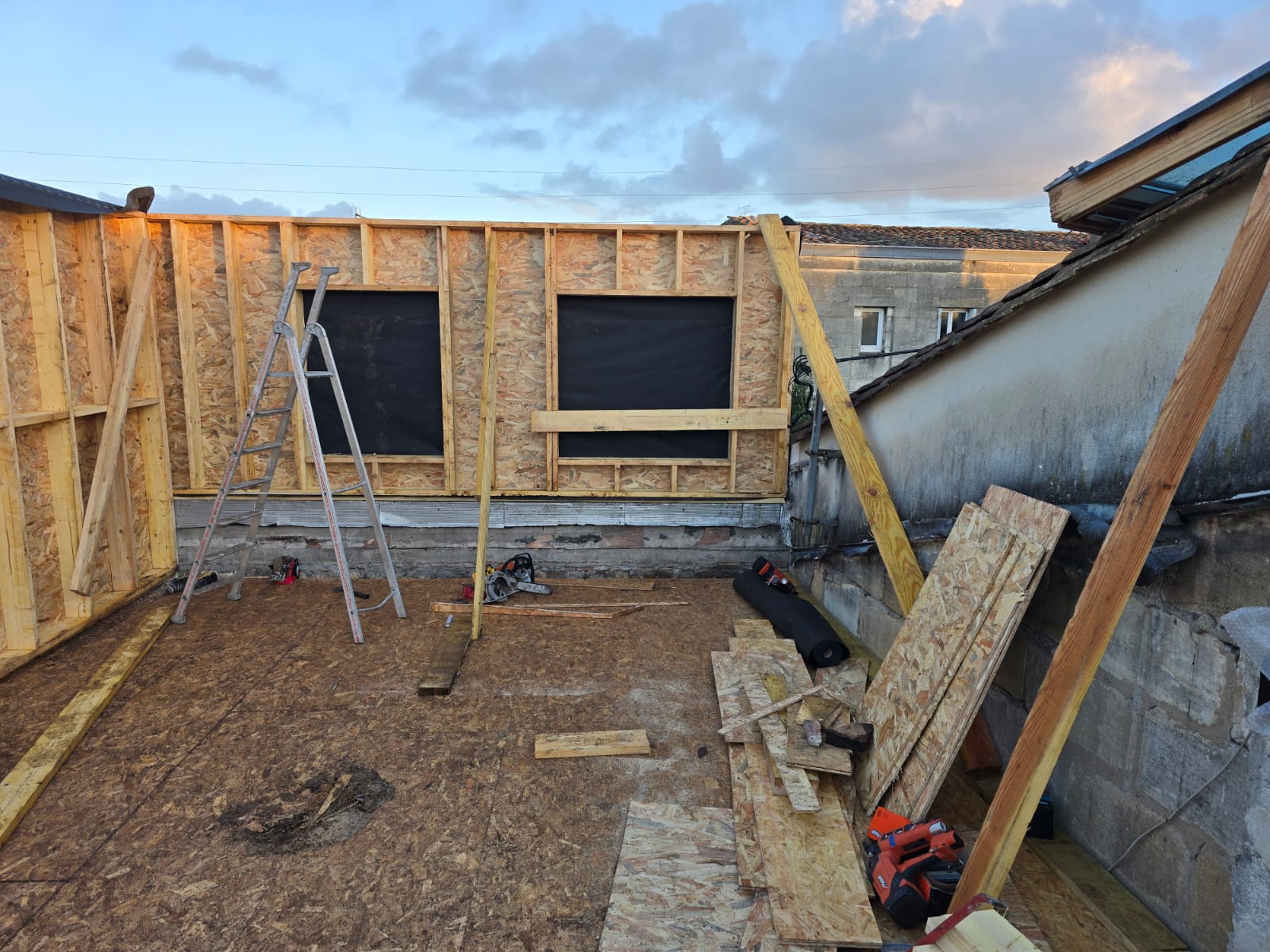 Chantier d'une extension en ossature bois avec murs OSB, échelle et outils de construction au coucher du soleil.