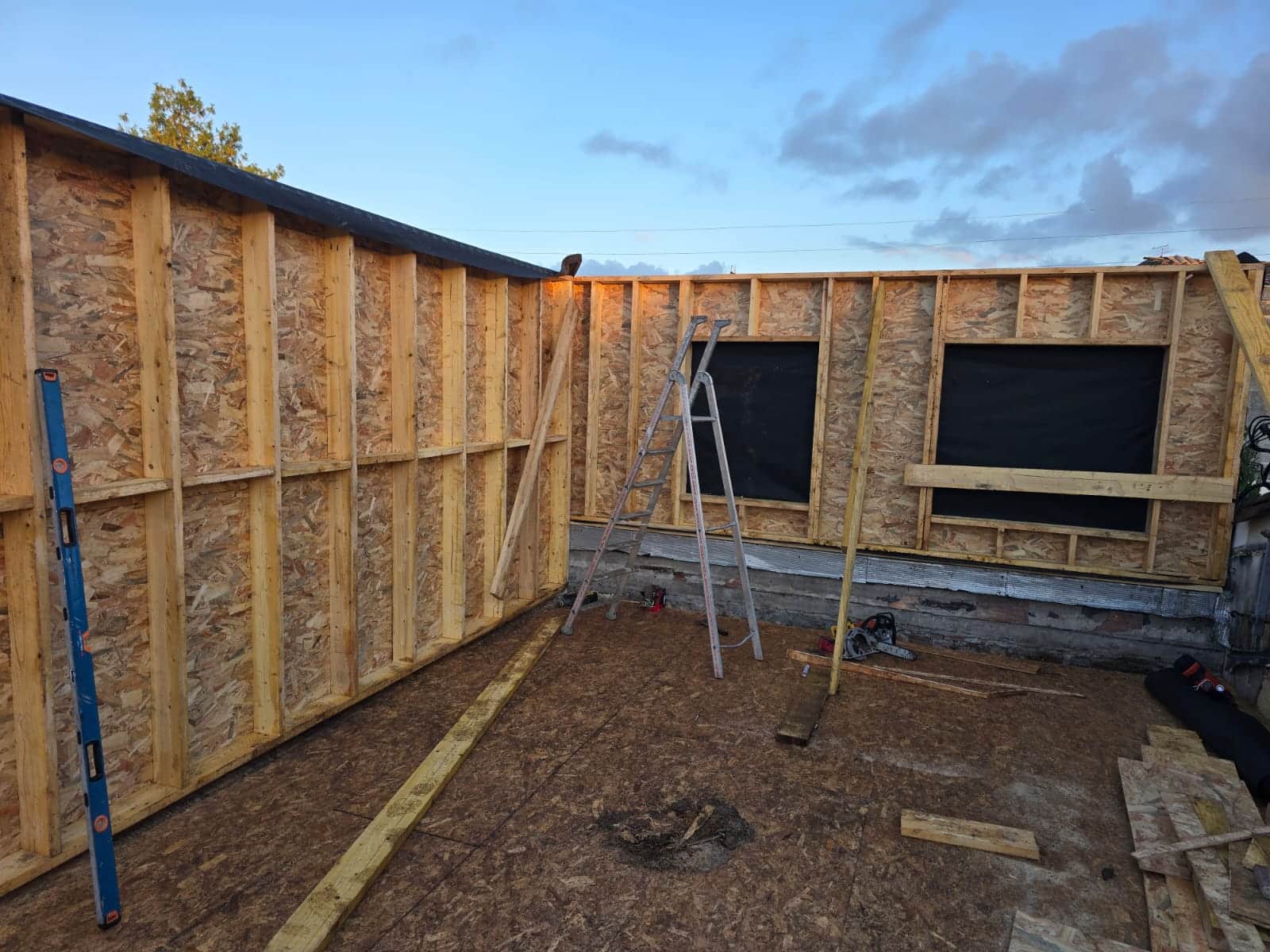 Construction d'une extension en bois avec panneaux OSB, charpente apparente, échelle et outils sur un chantier.
