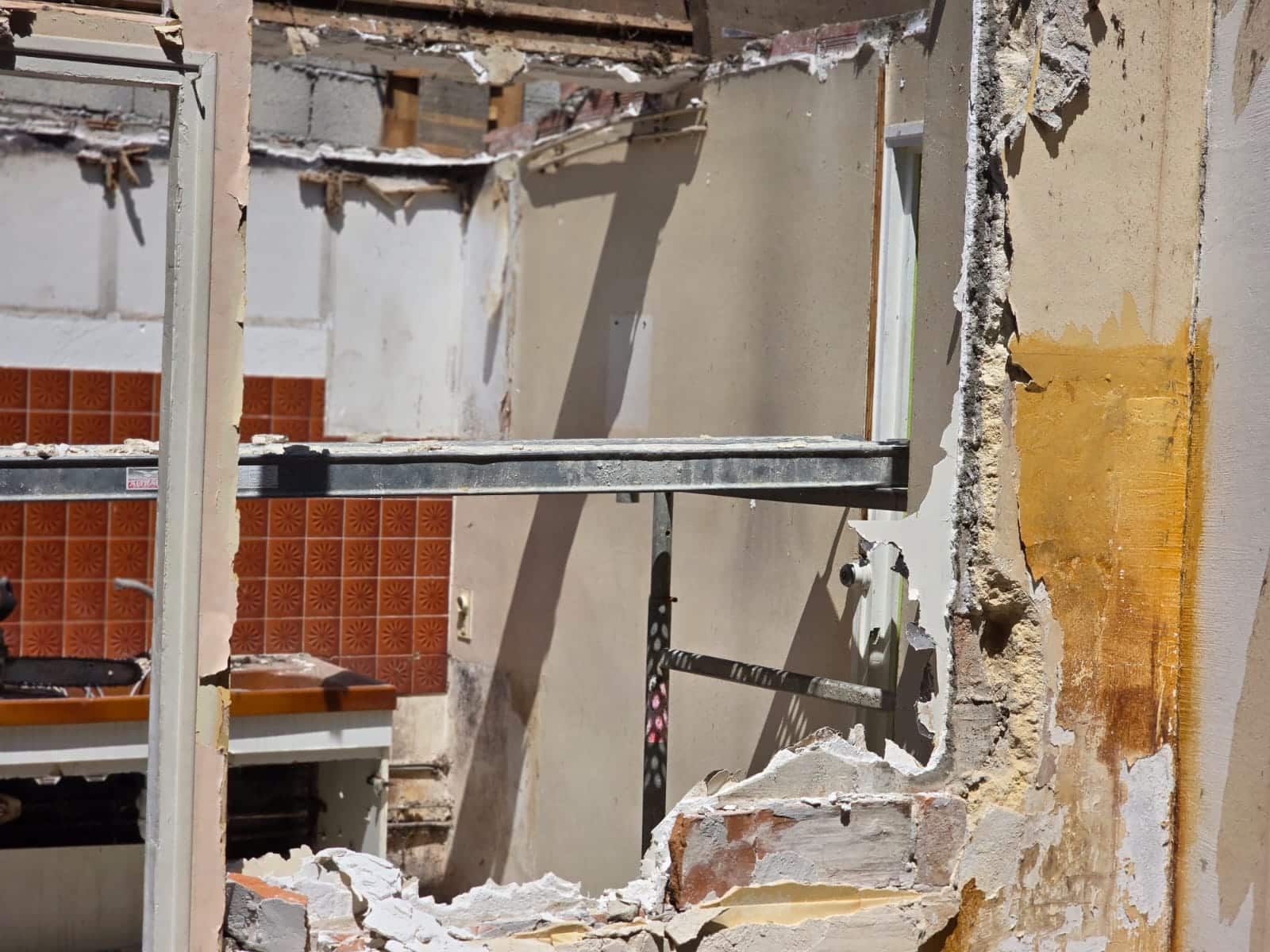 Maison en démolition avec murs brisés, carrelage orange et poutre d'échafaudage parmi les gravats.