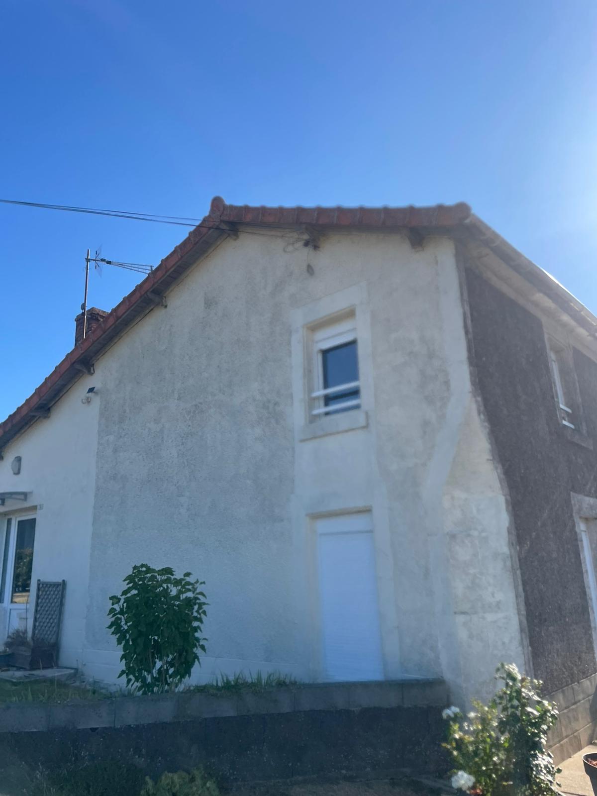 Façade latérale d'une maison ancienne avec crépi blanc, fenêtres blanches et toit en tuiles rouges sous un ciel bleu.