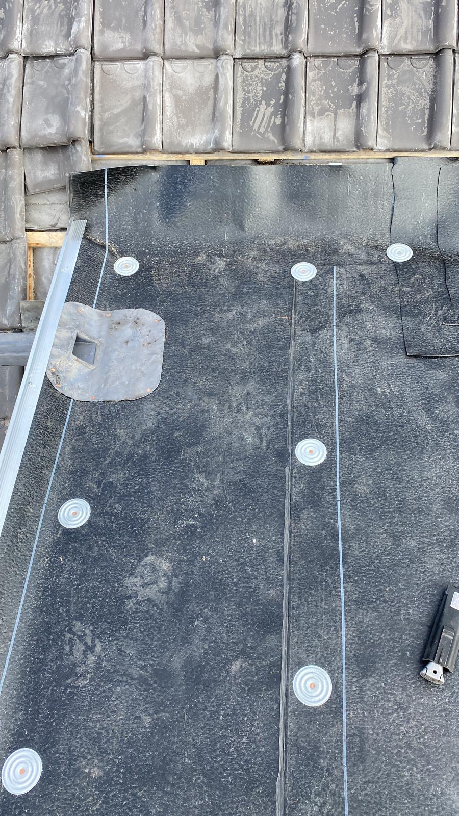 Installation d'une membrane d'étanchéité noire fixée par rondelles métalliques sur un toit avant la pose des tuiles.