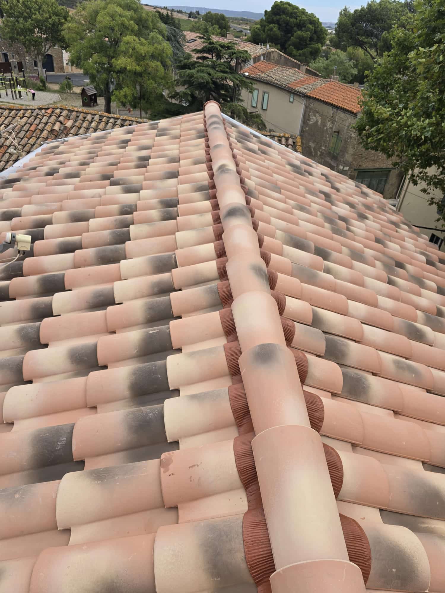 Toit en tuiles canal bicolores avec faîtage, dominant un village pittoresque et verdoyant.