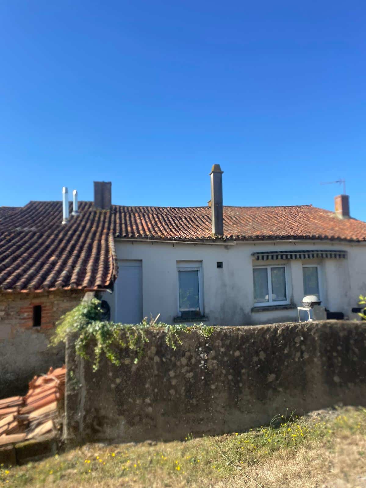 Maison blanche traditionnelle avec toit en tuiles, cheminées et muret de pierre sous un ciel bleu ensoleillé.