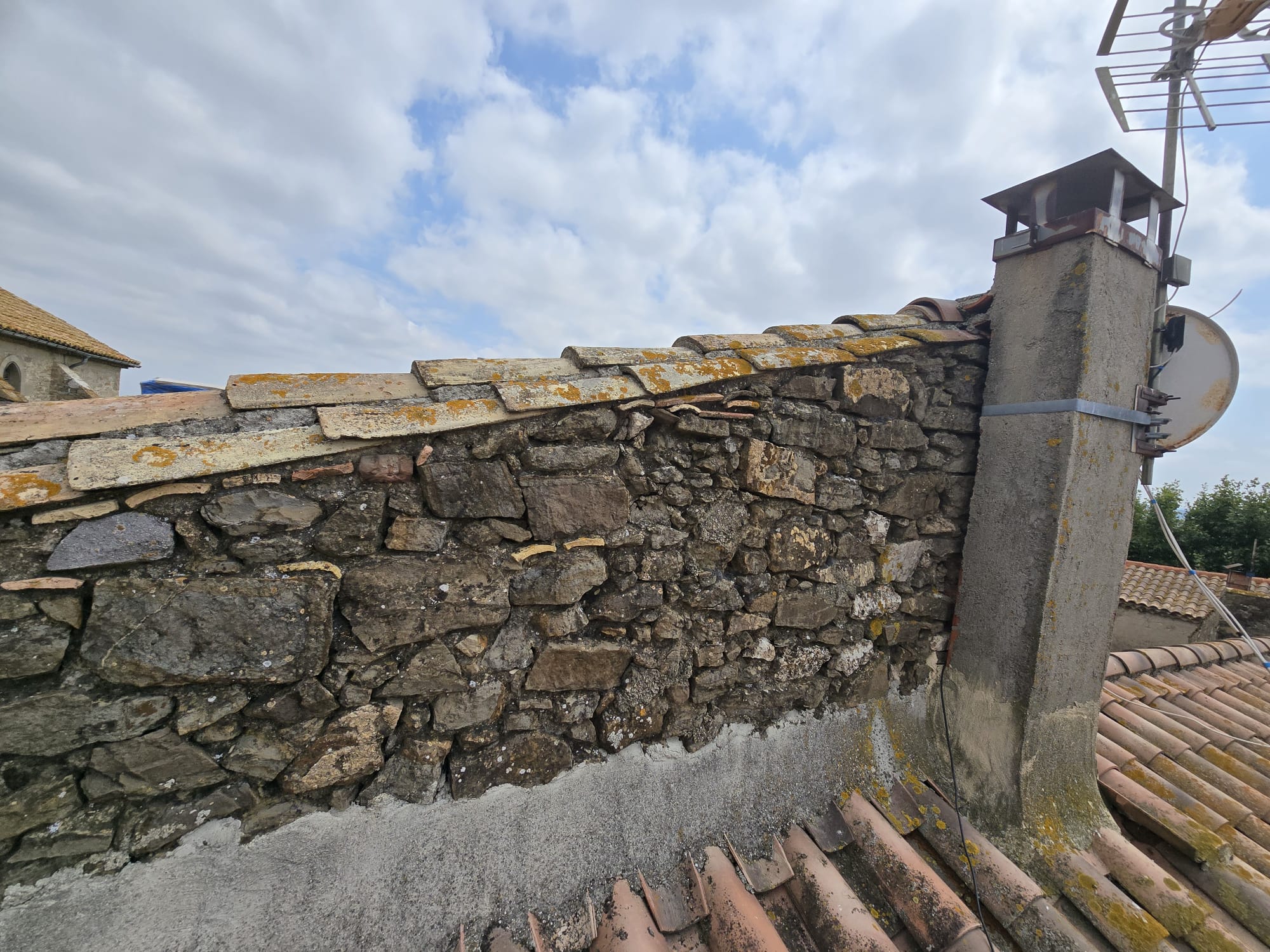 Mur en pierres apparentes sur toiture en tuiles avec cheminée et antenne sous un ciel bleu nuageux.