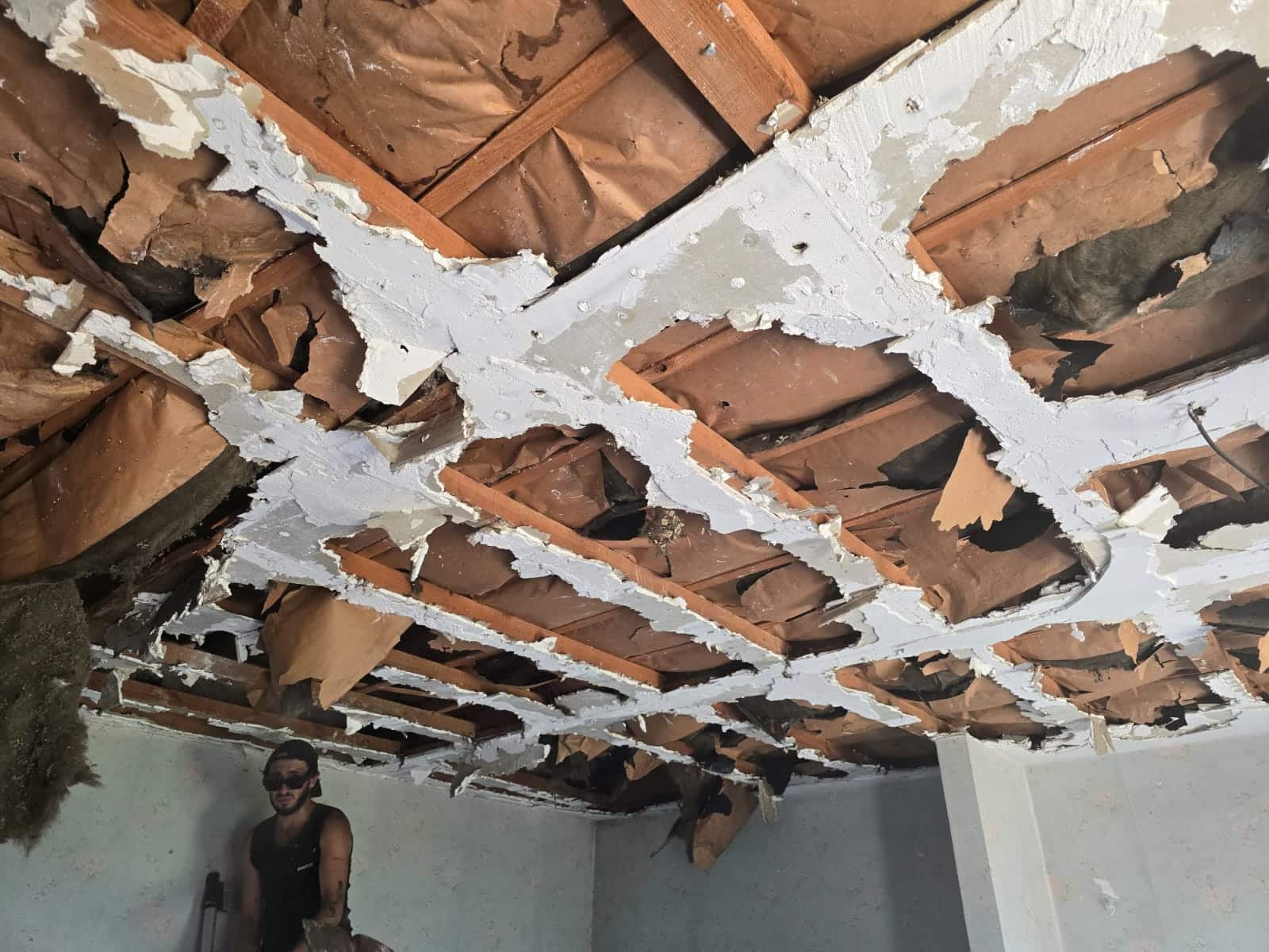 Plafond démoli avec solives en bois et isolant apparents, un homme en tenue de travail inspecte les dégâts.