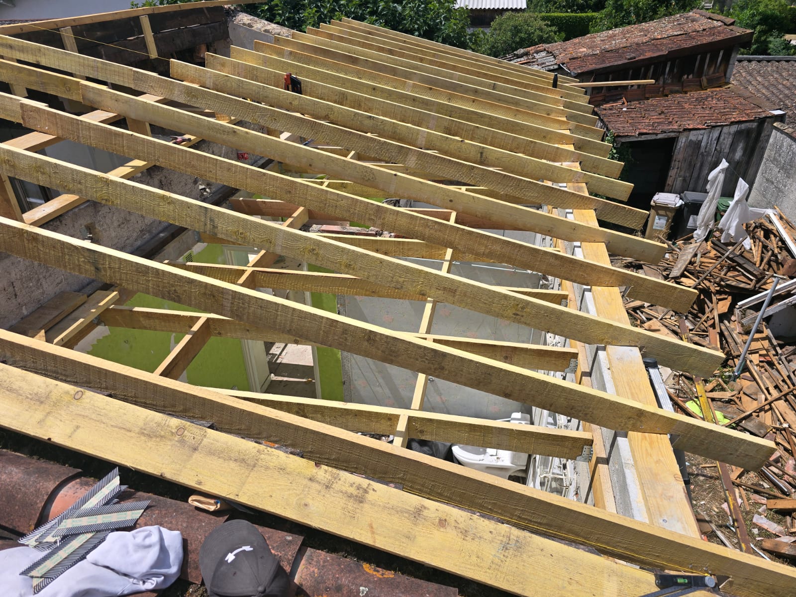Charpente de toit en bois en rénovation avec chevrons neufs au-dessus d'une salle de bain et débris extérieurs.