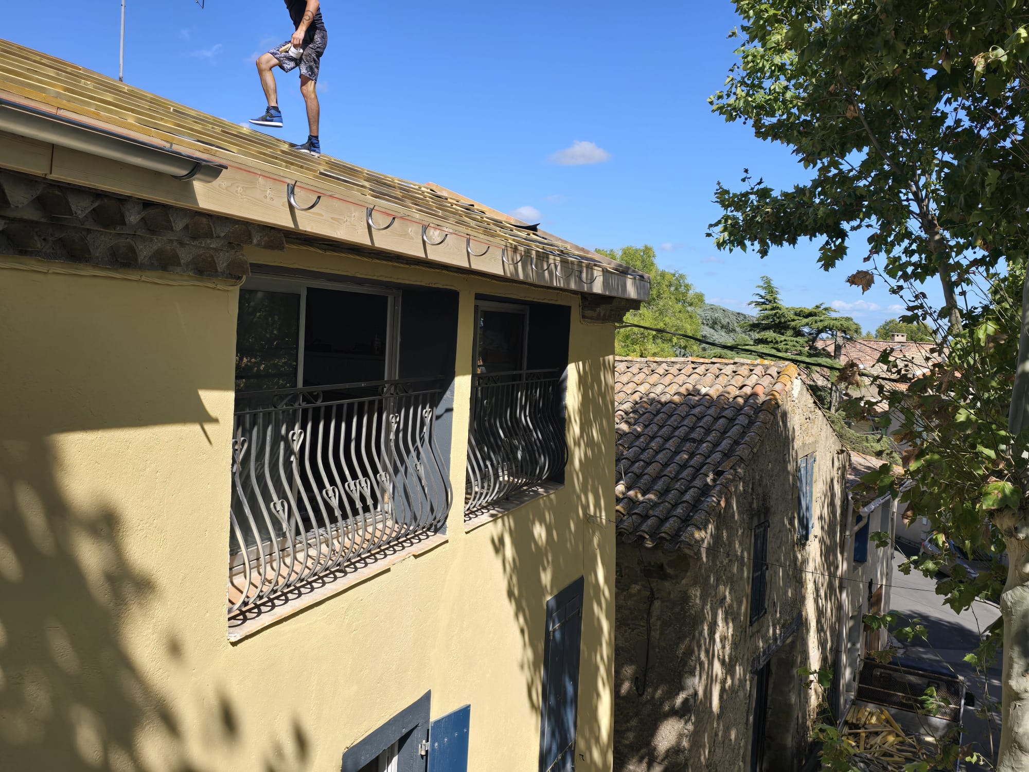 Couvreur sur le toit d'une maison provençale jaune en rénovation, installation de gouttières et charpente apparente.
