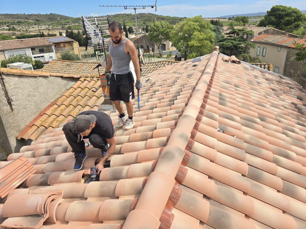 Deux couvreurs rénovent un toit en tuiles romanes. Un ouvrier utilise une perceuse électrique sur une tuile.