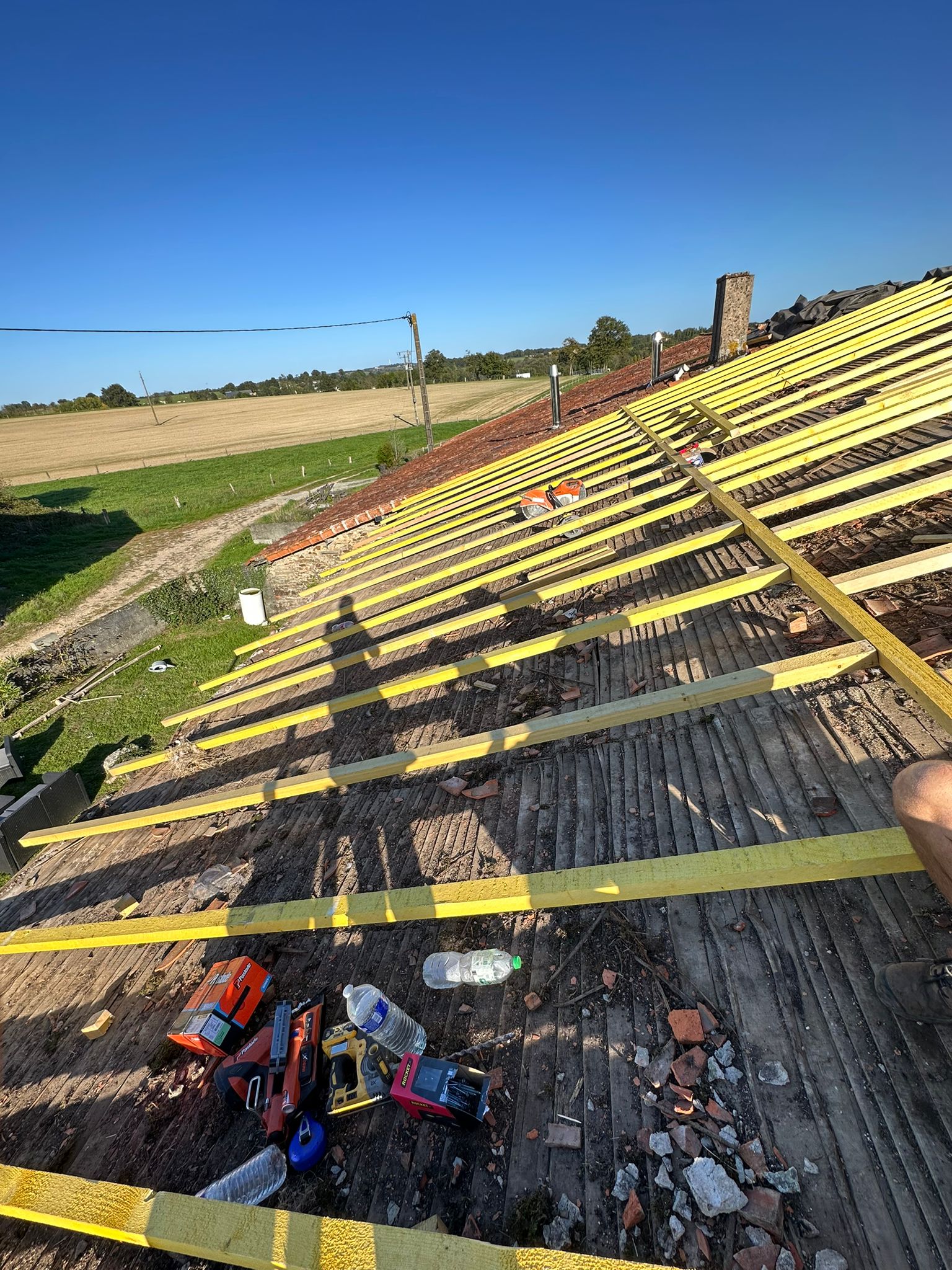 Pose de liteaux en bois jaune sur une charpente ancienne en rénovation avec des outils de couvreur au soleil.