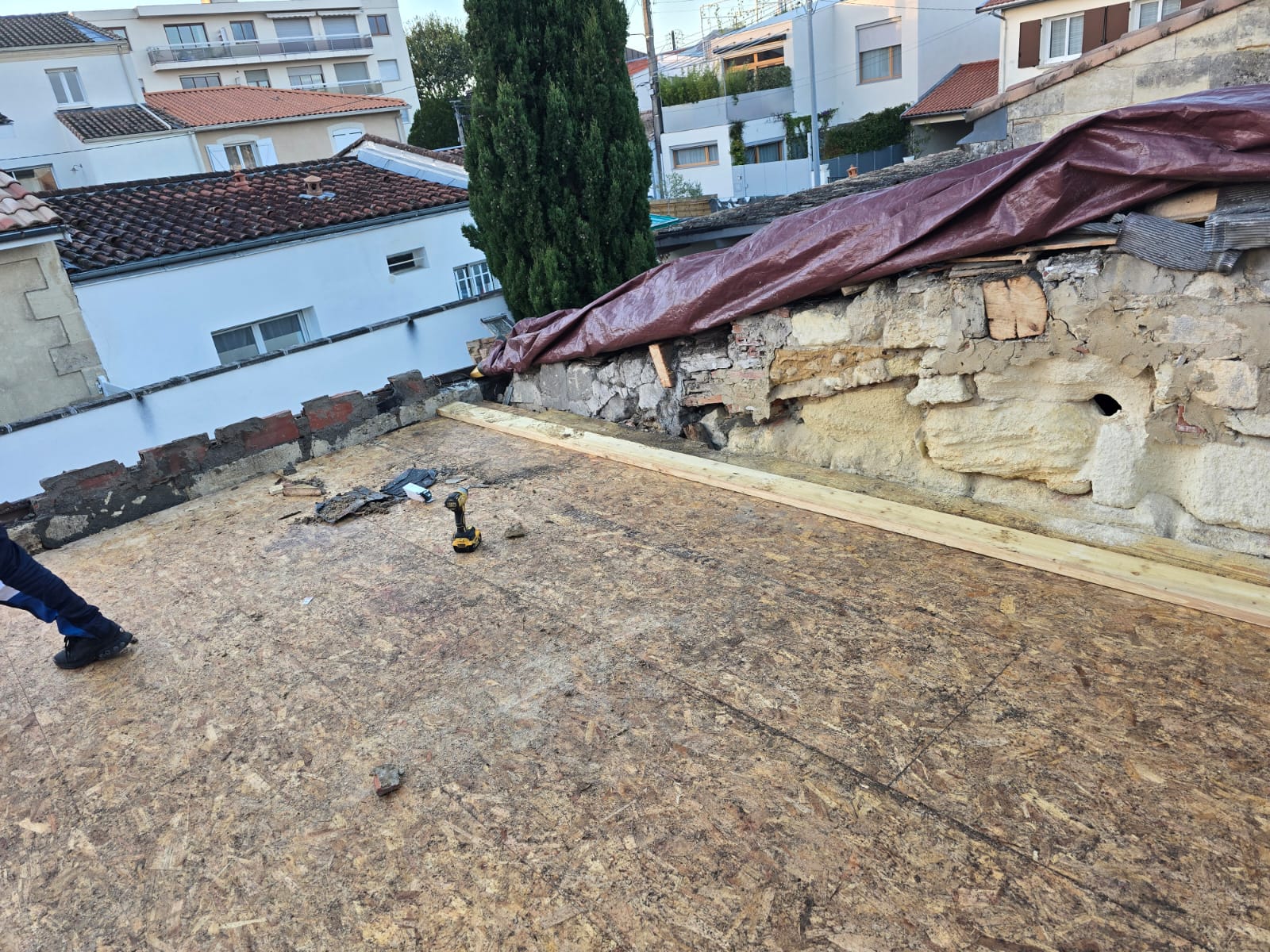 Panneaux OSB installés sur un toit plat en rénovation, bordé d'un mur en pierre et d'une bâche de protection.