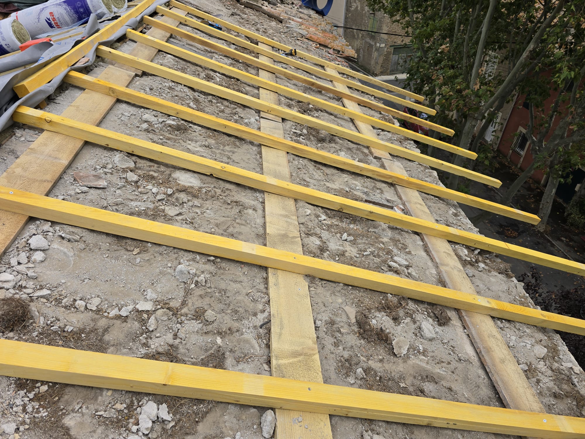 Pose de liteaux en bois jaune sur une toiture en rénovation avec isolant URSA visible en arrière-plan.