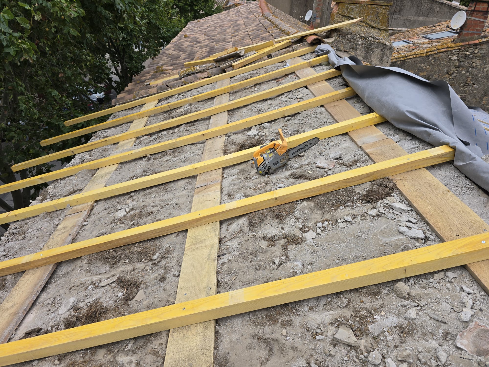 Rénovation de toiture avec nouveaux liteaux en bois et une tronçonneuse posée sur les gravats du chantier.