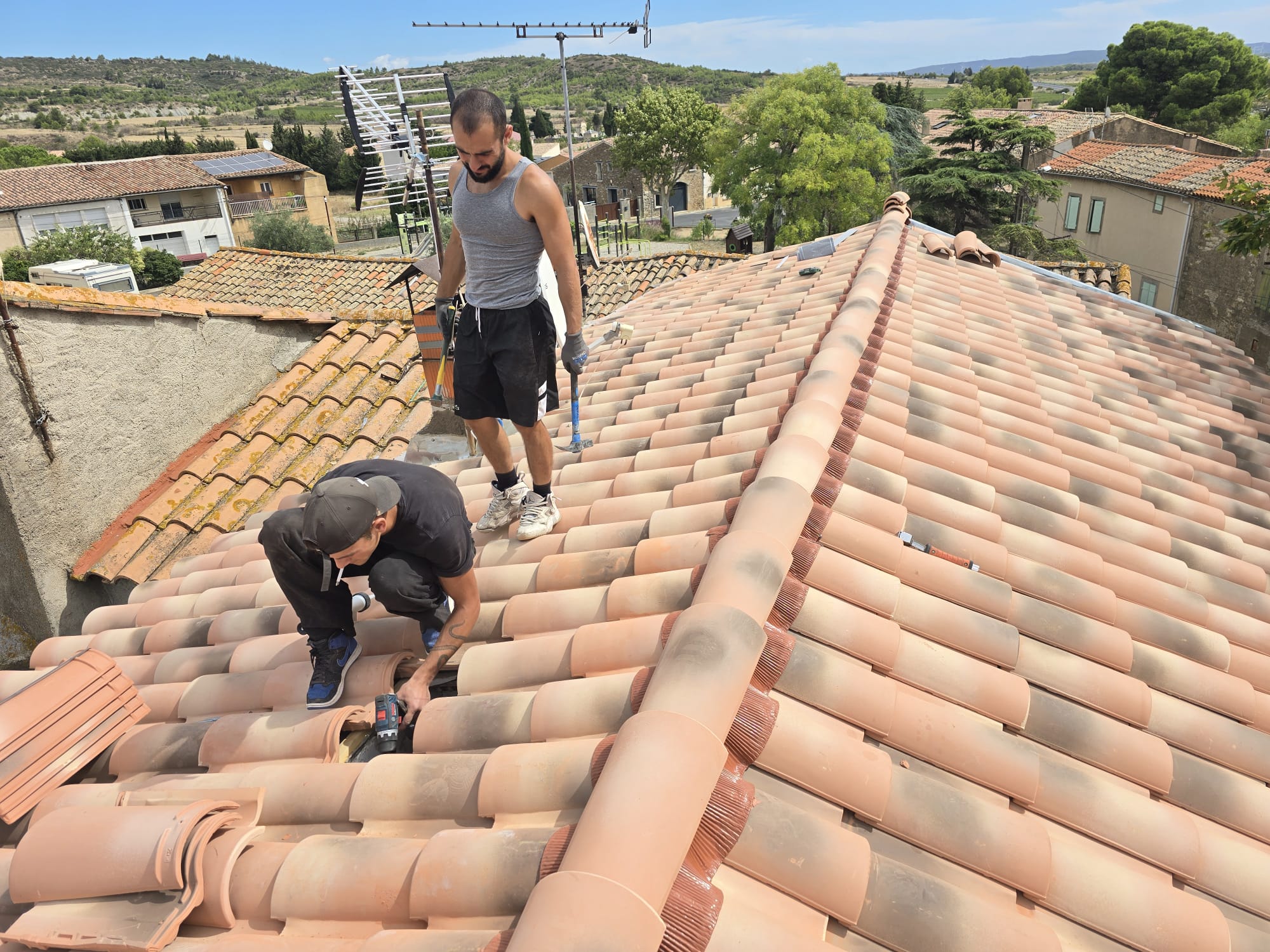 Deux artisans couvreurs travaillent sur la rénovation d'un toit en tuiles canal sous un soleil radieux.