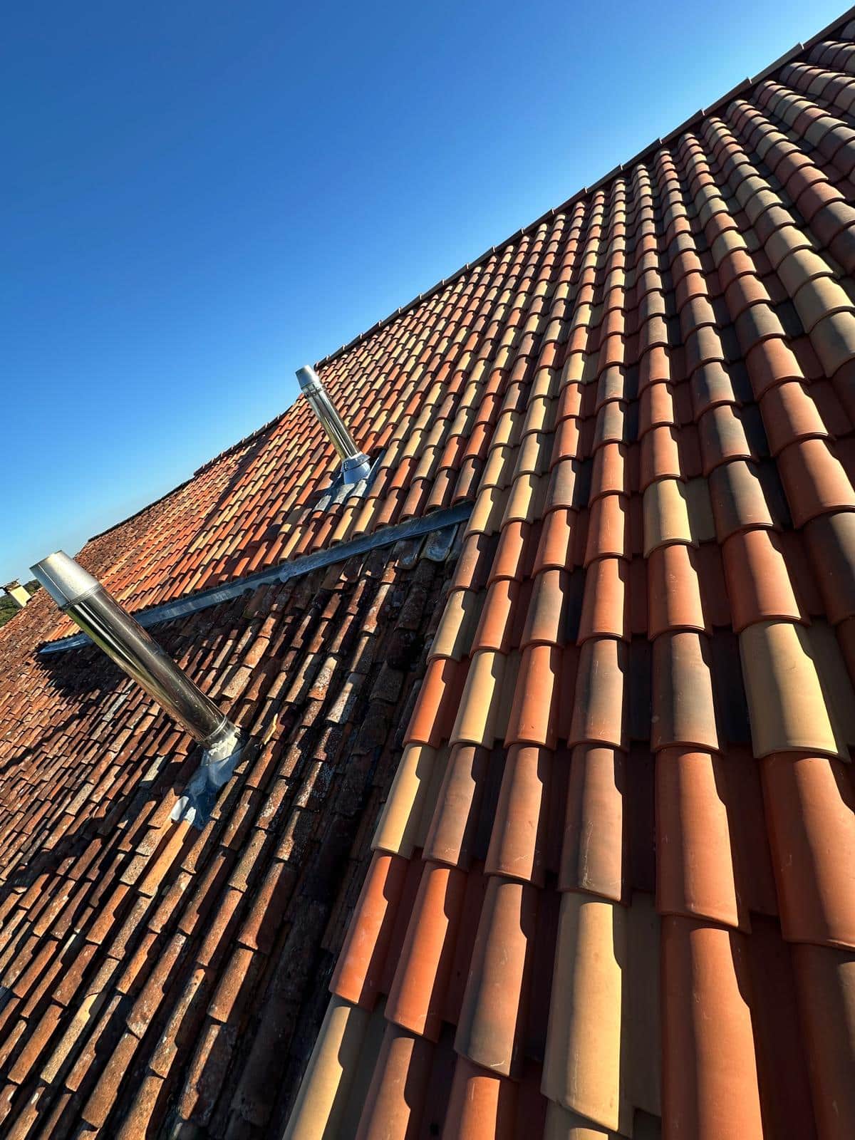 Toiture en tuiles canal mêlant tuiles anciennes et neuves avec deux conduits de cheminée en inox sous ciel bleu.