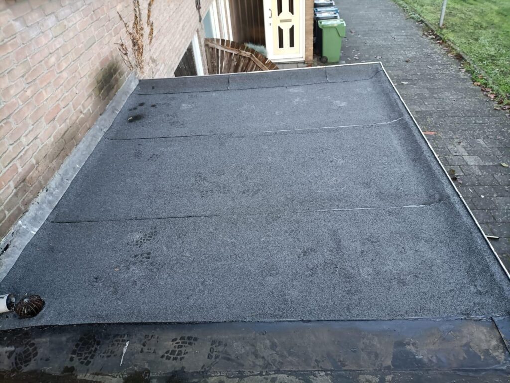 Toiture plate en membrane bitumineuse grise avec système d'évacuation, bordant un mur de briques extérieur.