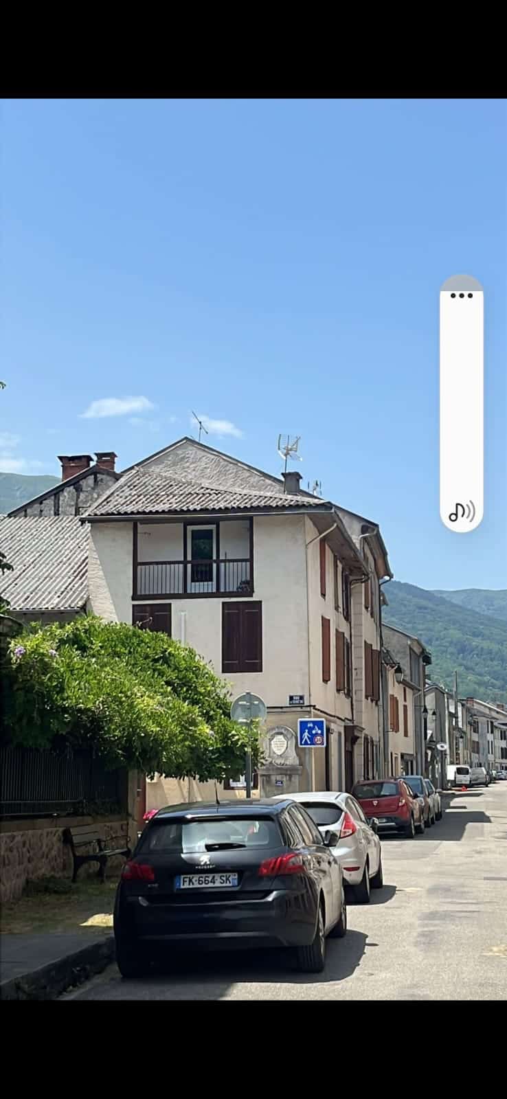 Rue de village en montagne avec maisons traditionnelles, voitures garées dont une Peugeot FK-664-SK sous un ciel bleu.